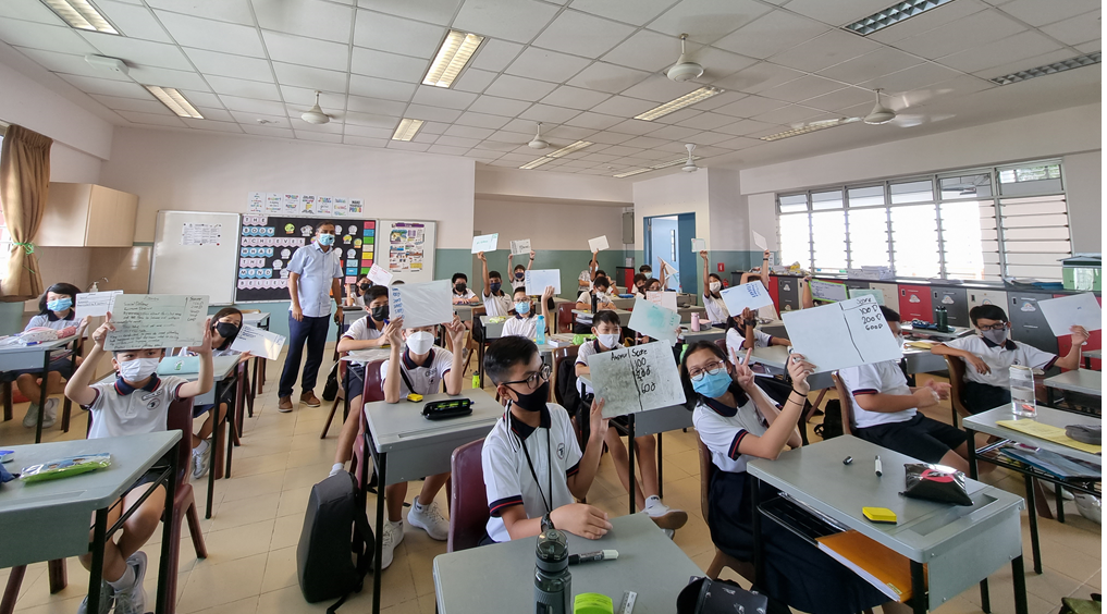 Students wearing masks sit at desks in a classroom, holding up papers with writing toward the camera.